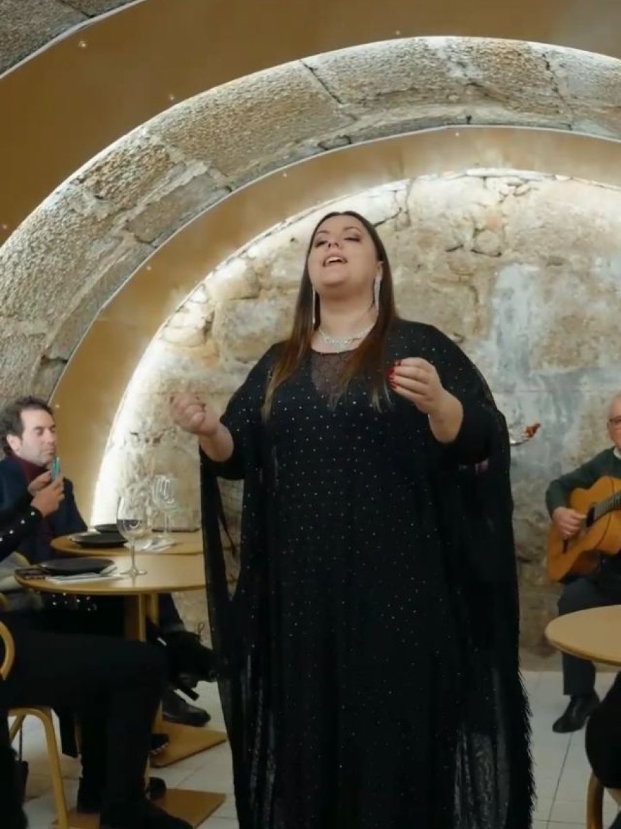 Fadista de xaile preto a cantar sob o arco de pedra da sala, com o guitarrista sentado ao lado e as mesas ocupadas